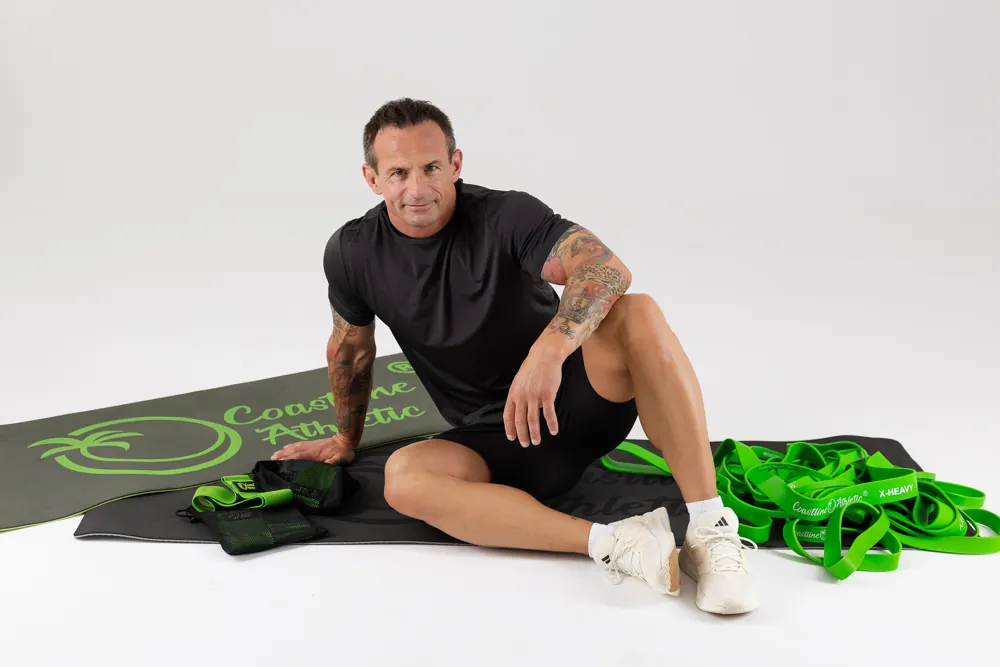 Mężczyzna siedzi na macie Coastline Athletic obok zielonych gum oporowych